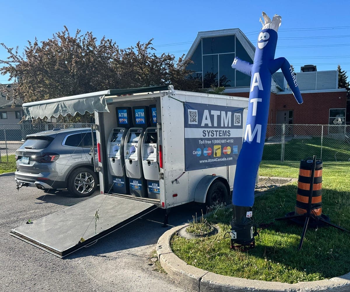 White ATM Systems trailer with rear ramp down holding three ATM machines, with a blue ATM air dancer beside it